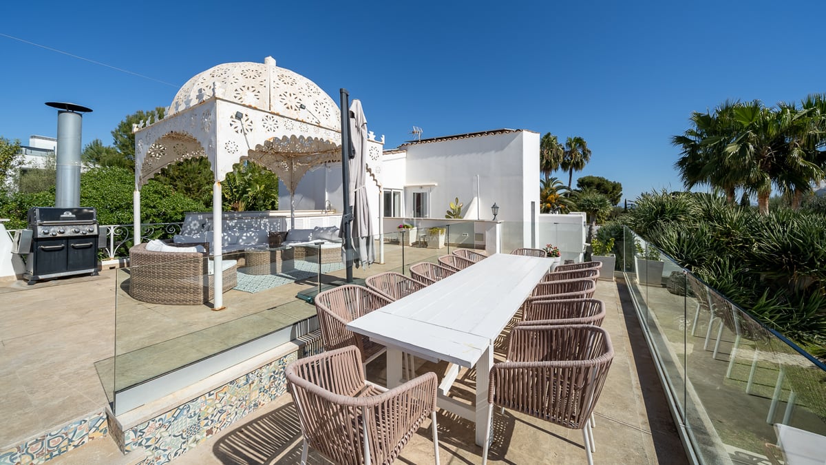 Grande terrasse avec table à manger blanche, chaises en osier, barbecue et gazebo orné sous un ciel bleu clair.
