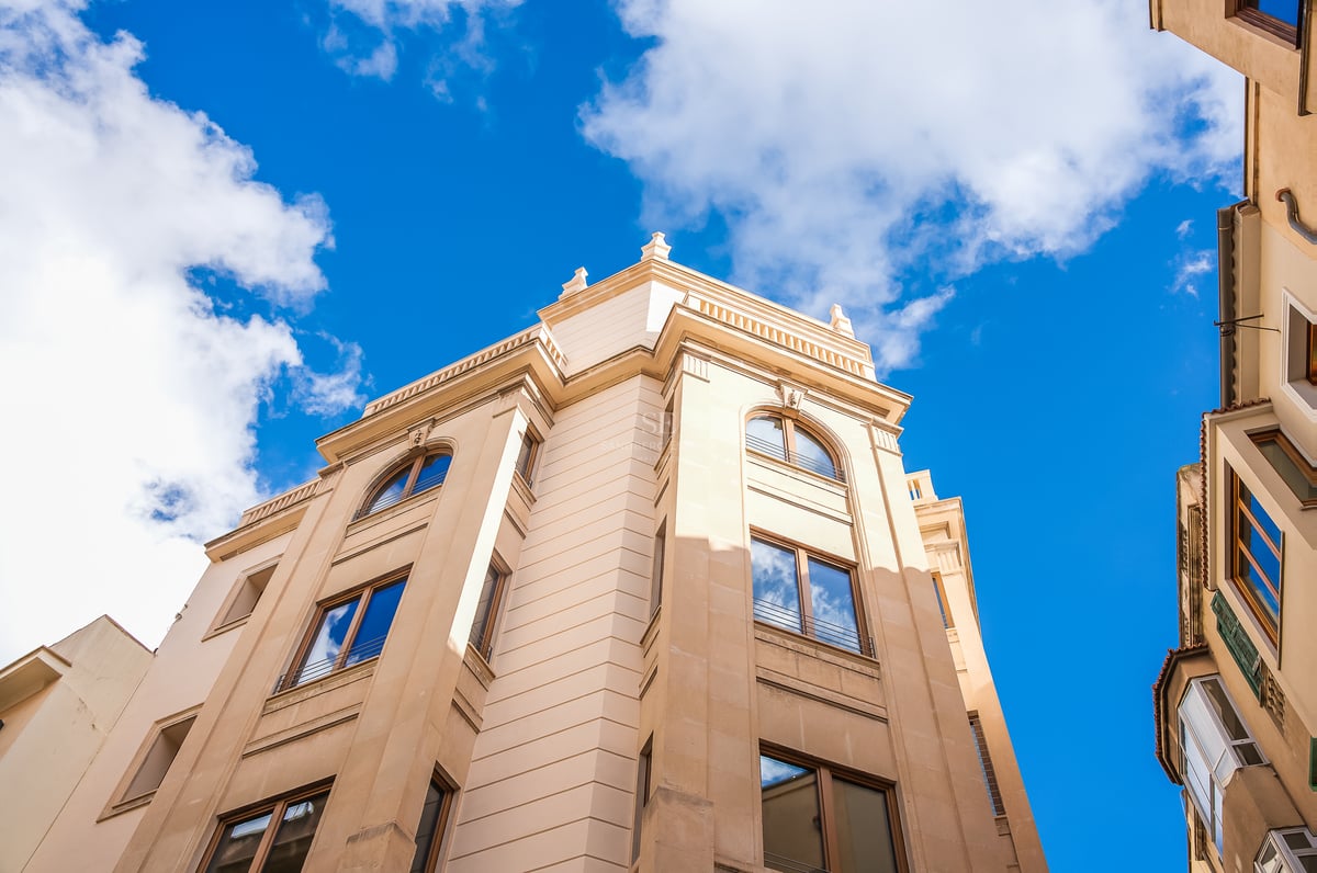 Low-angle view of a beige natural stone building facade with wooden window frames against a clear blue sky.