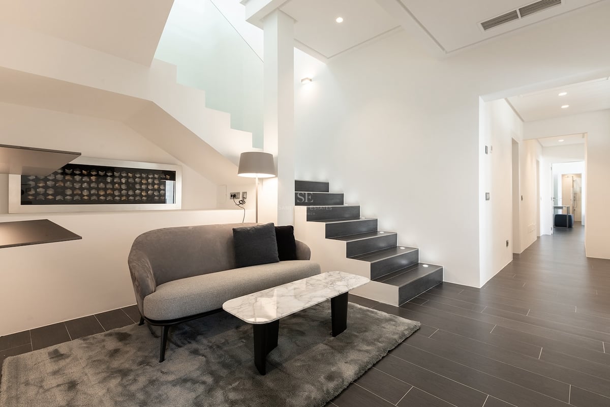 Espace salon moderne avec canapé gris, table en marbre et un escalier contemporain aux marches sombres avec éclairage intégré.