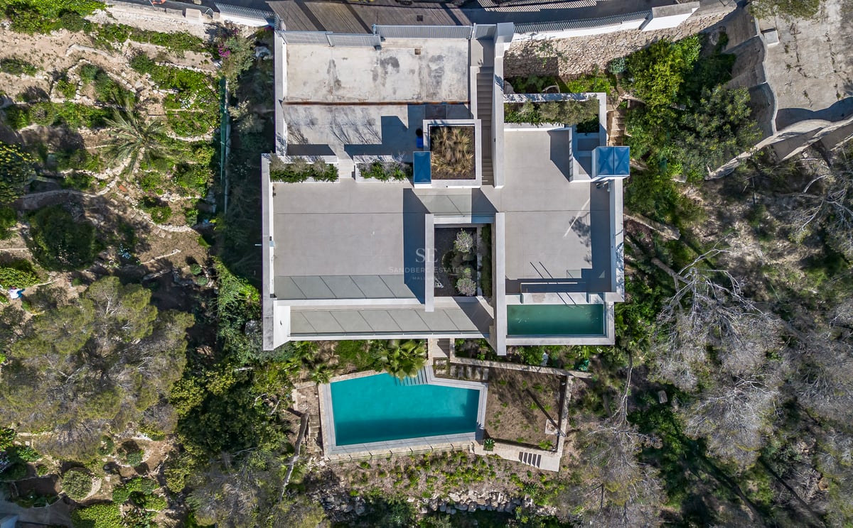 Vue aérienne d'une villa moderne géométrique avec toits plats, terrasses et piscine turquoise cristalline.