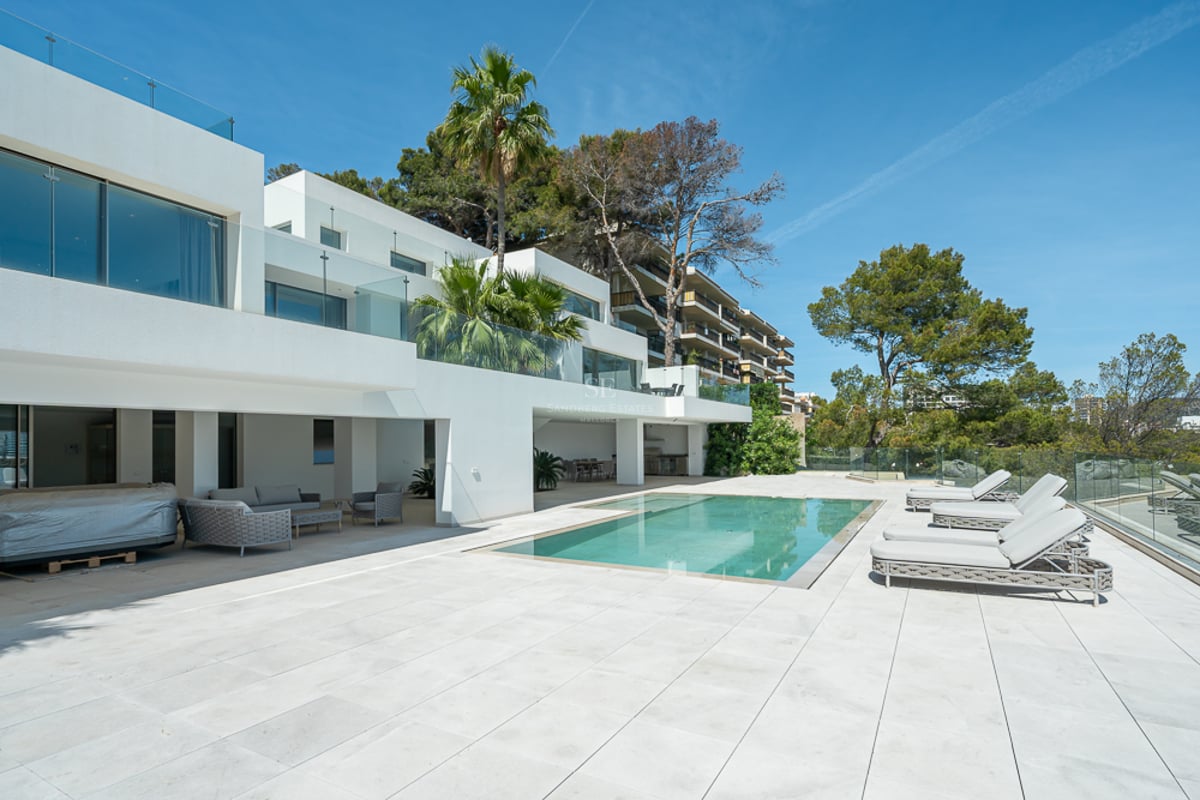 Villa blanche moderne avec piscine rectangulaire, bains de soleil gris et garde-corps en verre sous un ciel bleu.
