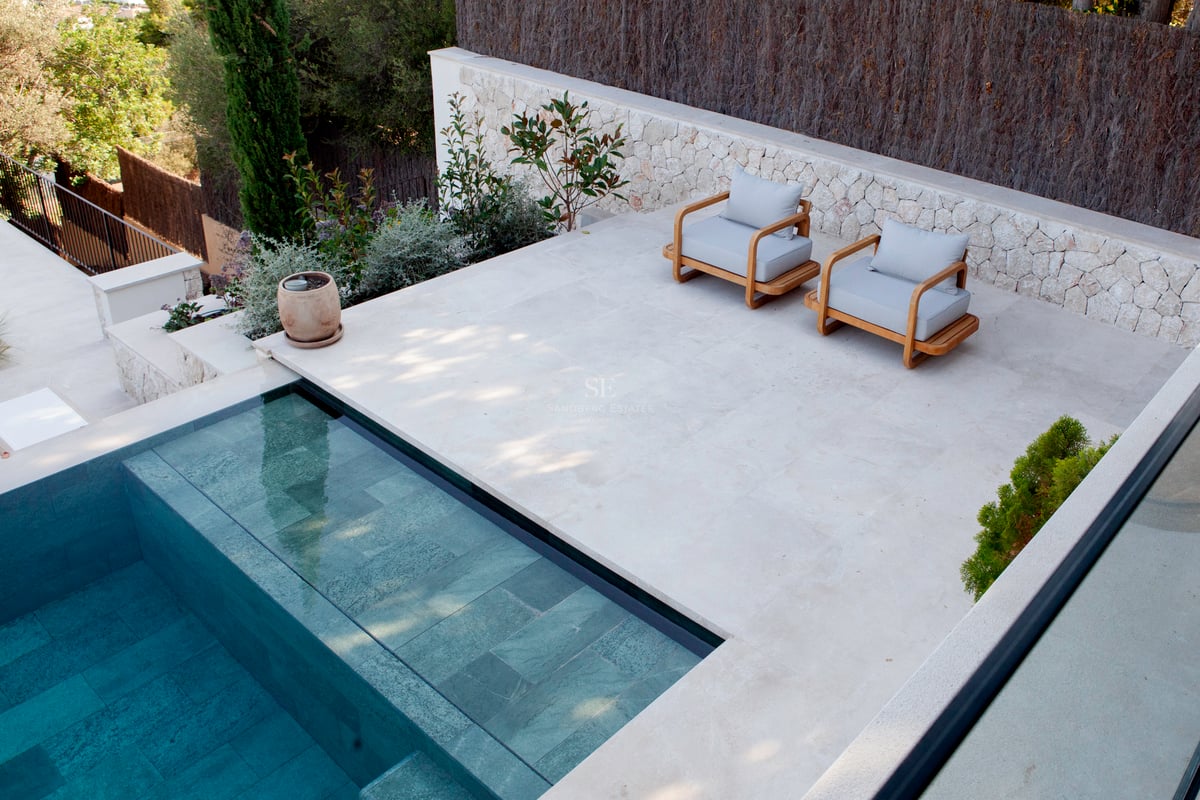 Vue en surplomb d'une piscine carrelée turquoise bordée d'une terrasse en pierre claire avec fauteuils en bois.