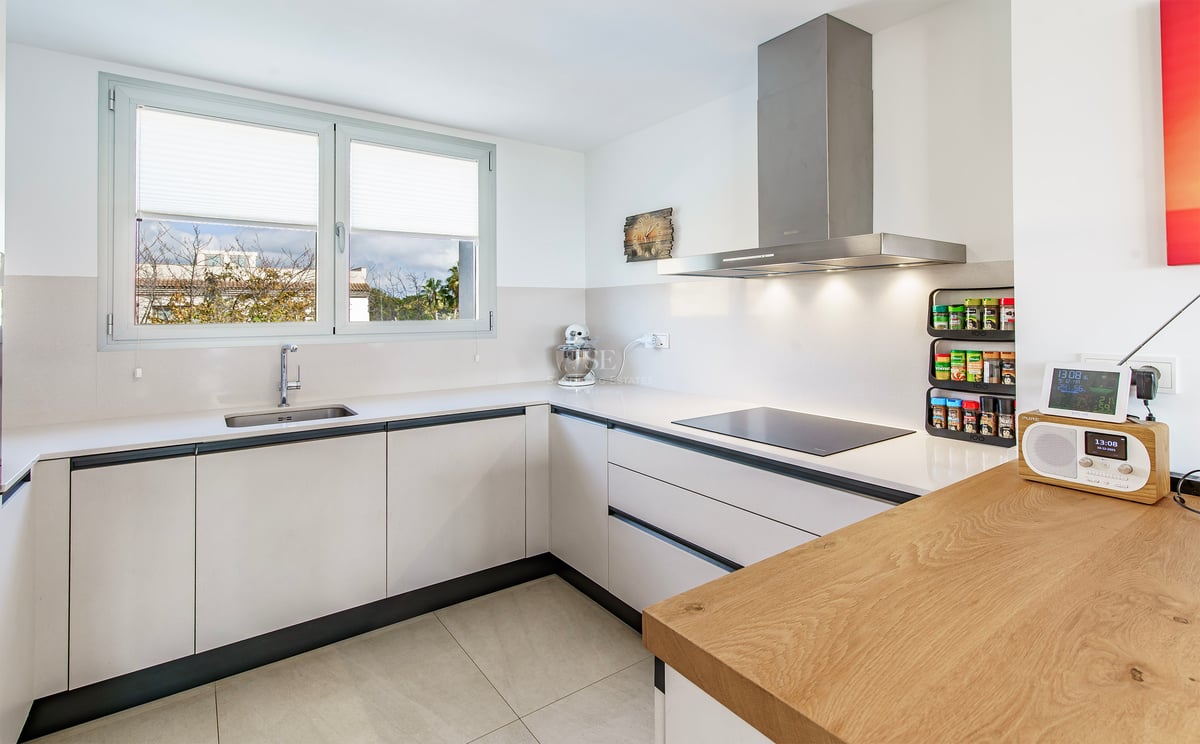 Cuisine blanche moderne en L avec plan de travail en bois, hotte en inox et grande fenêtre.