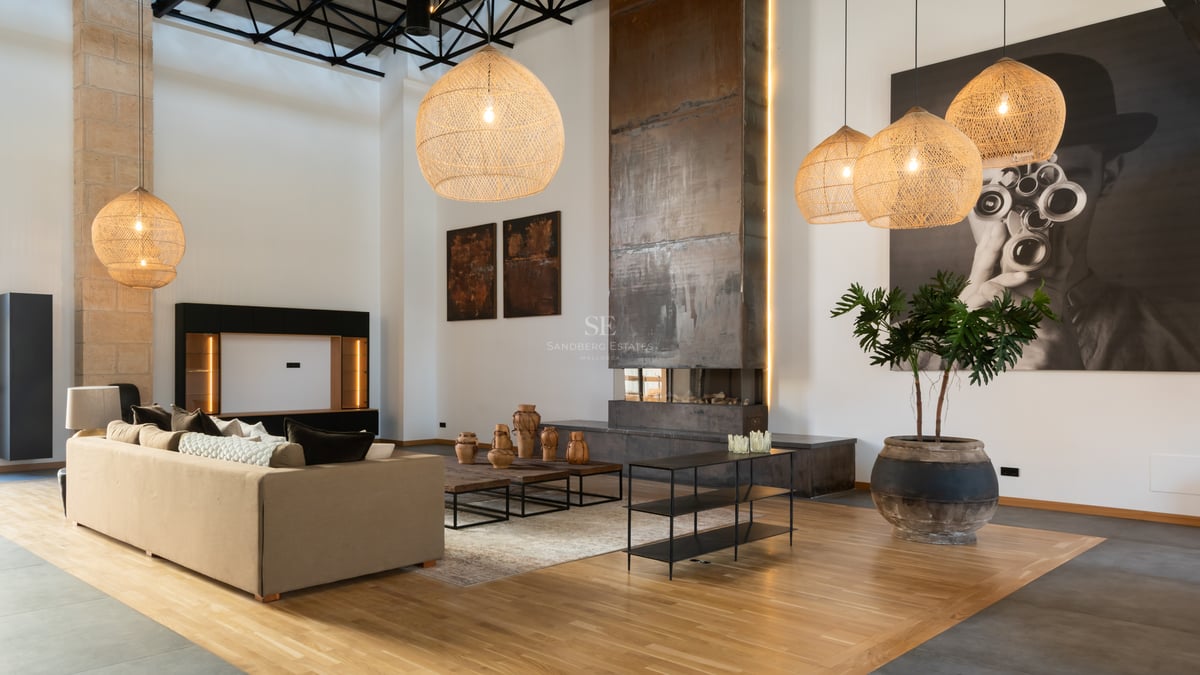 Grand salon avec de hauts plafonds, une immense cheminée en métal, des suspensions en osier et un canapé beige.