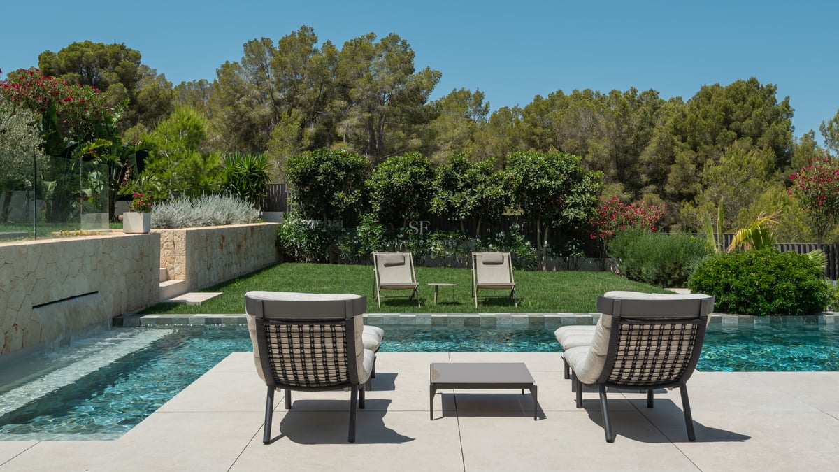 Espace piscine avec bains de soleil modernes, pelouse soignée et mur en pierre avec cascade sous un ciel bleu.