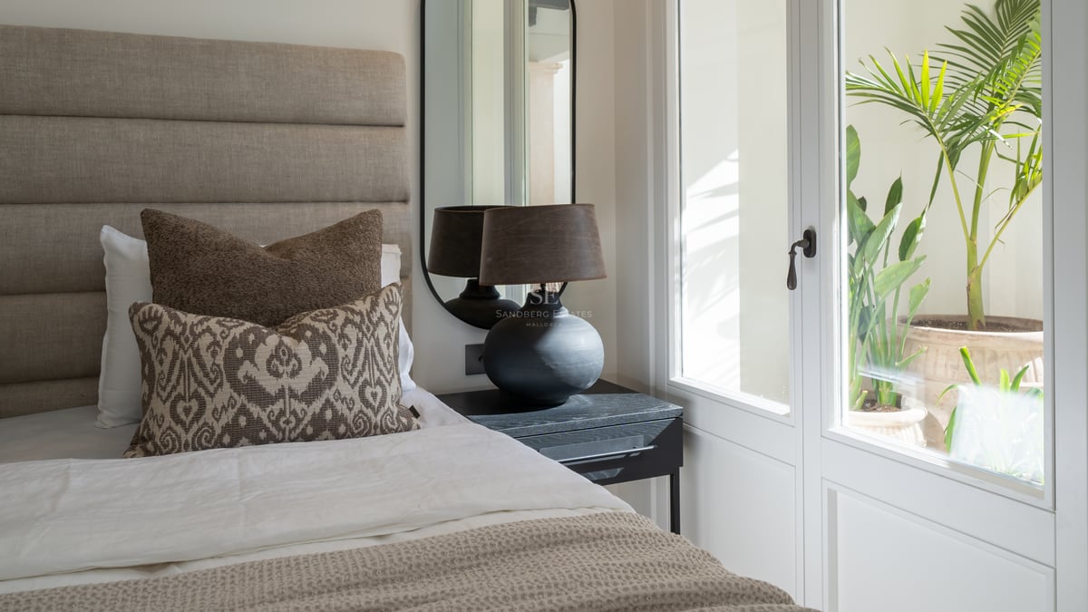 Contemporary bedroom interior with a linen headboard, black designer lamp, and large window overlooking a lush courtyard.