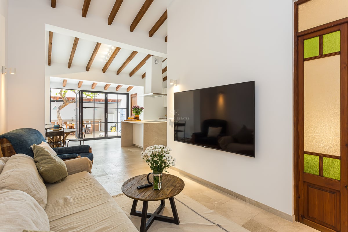 Salon moderne avec canapé beige, poutres en bois au plafond, grand téléviseur et vue sur la cuisine ouverte.