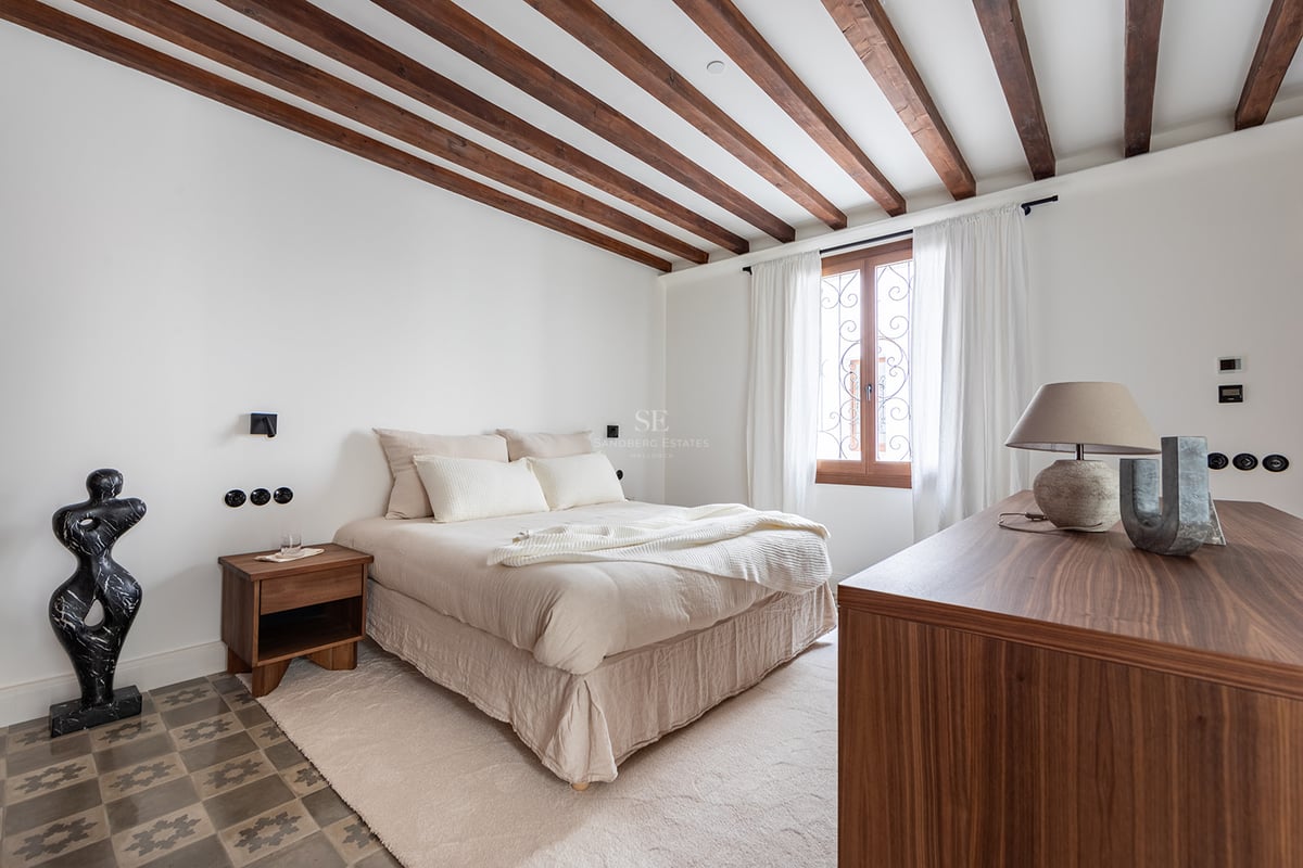 Dormitorio con vigas de madera oscura, suelo de baldosas hidráulicas y una ventana con cortinas blancas.