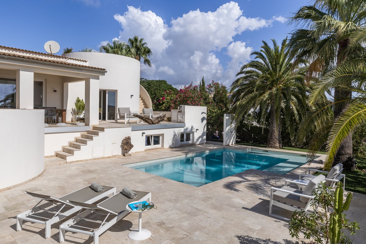 Villa blanche moderne avec piscine turquoise, terrasse en pierre naturelle et palmiers sous un ciel bleu éclatant.