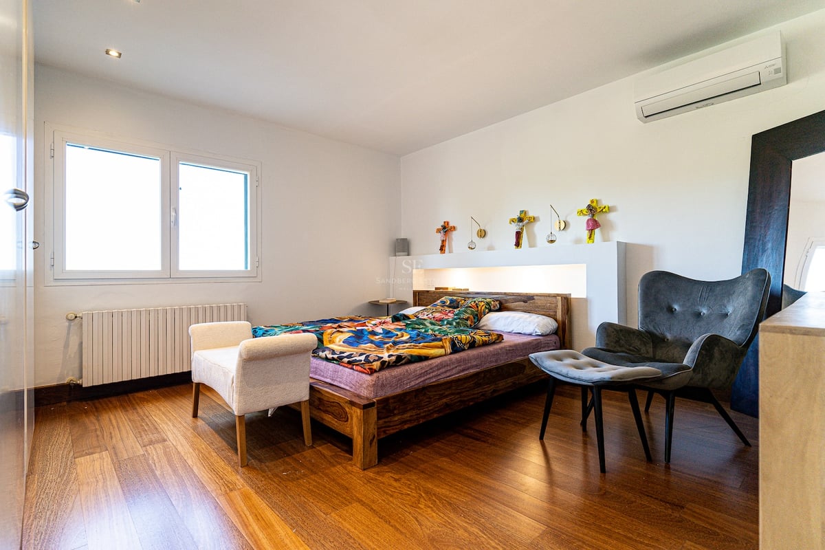 Chambre lumineuse avec parquet, cadre de lit en bois, fauteuil gris et fenêtre laissant entrer la lumière naturelle.