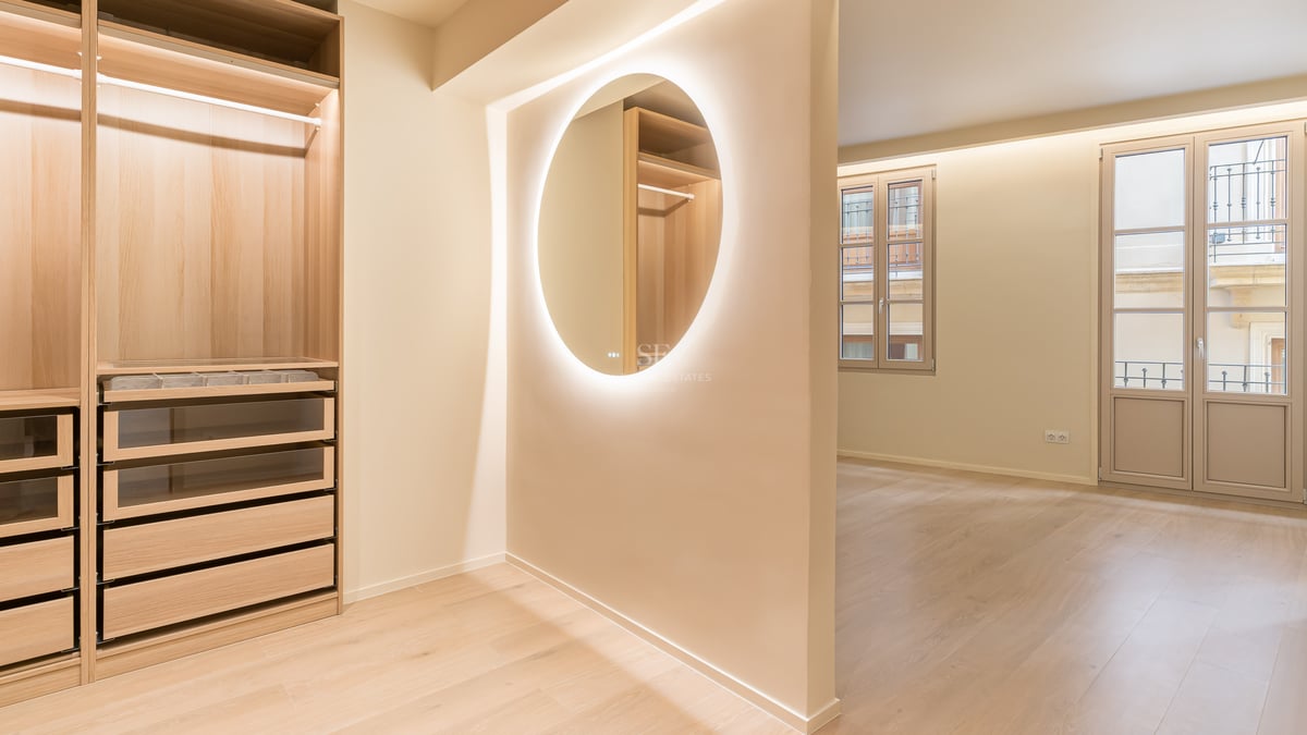Luxury walk-in dressing area with light oak wardrobes, backlit circular mirror, and glass-fronted drawers.