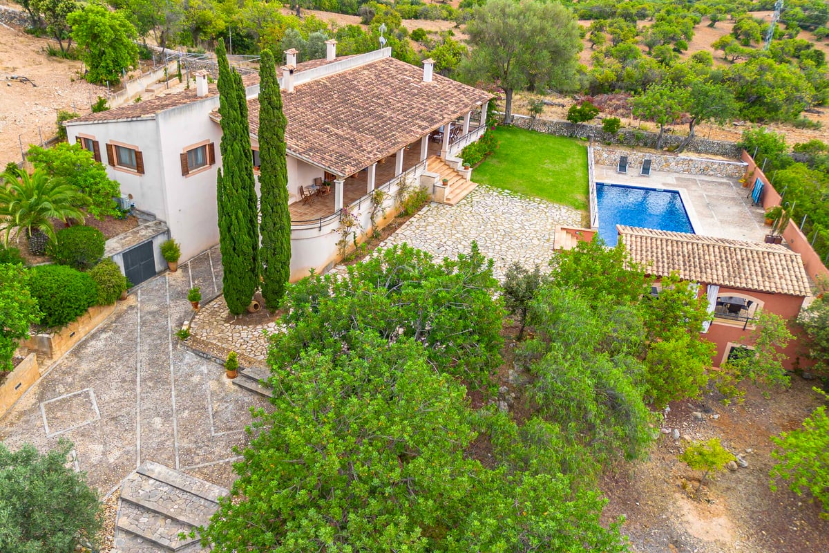 Vue en plongée d'une villa avec toit en terre cuite, cour en pierre, piscine bleue et aménagement paysager verdoyant.