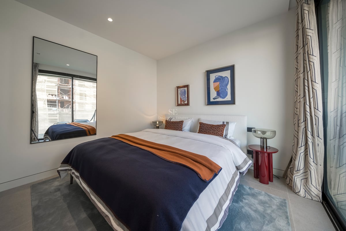 Chambre moderne lumineuse avec grand lit, literie bleu et orange, table rouge et un miroir reflétant une fenêtre.