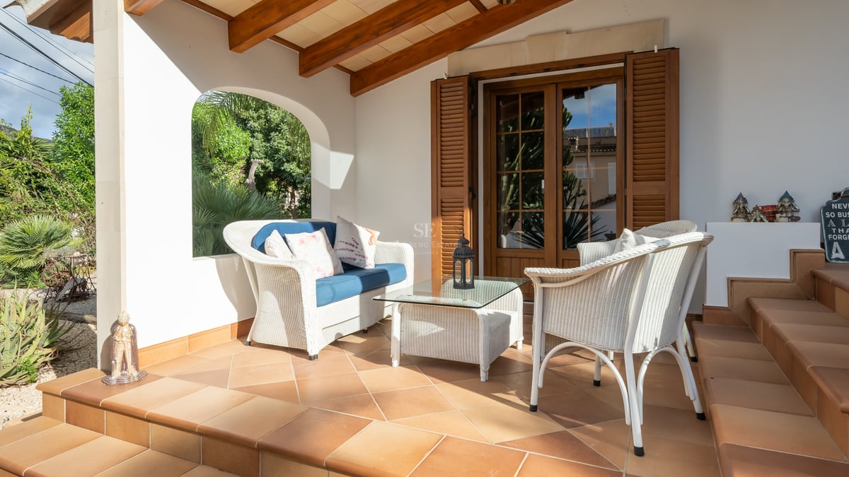 Mobilier en osier blanc sur une terrasse en terre cuite avec poutres en bois et vue sur le jardin.