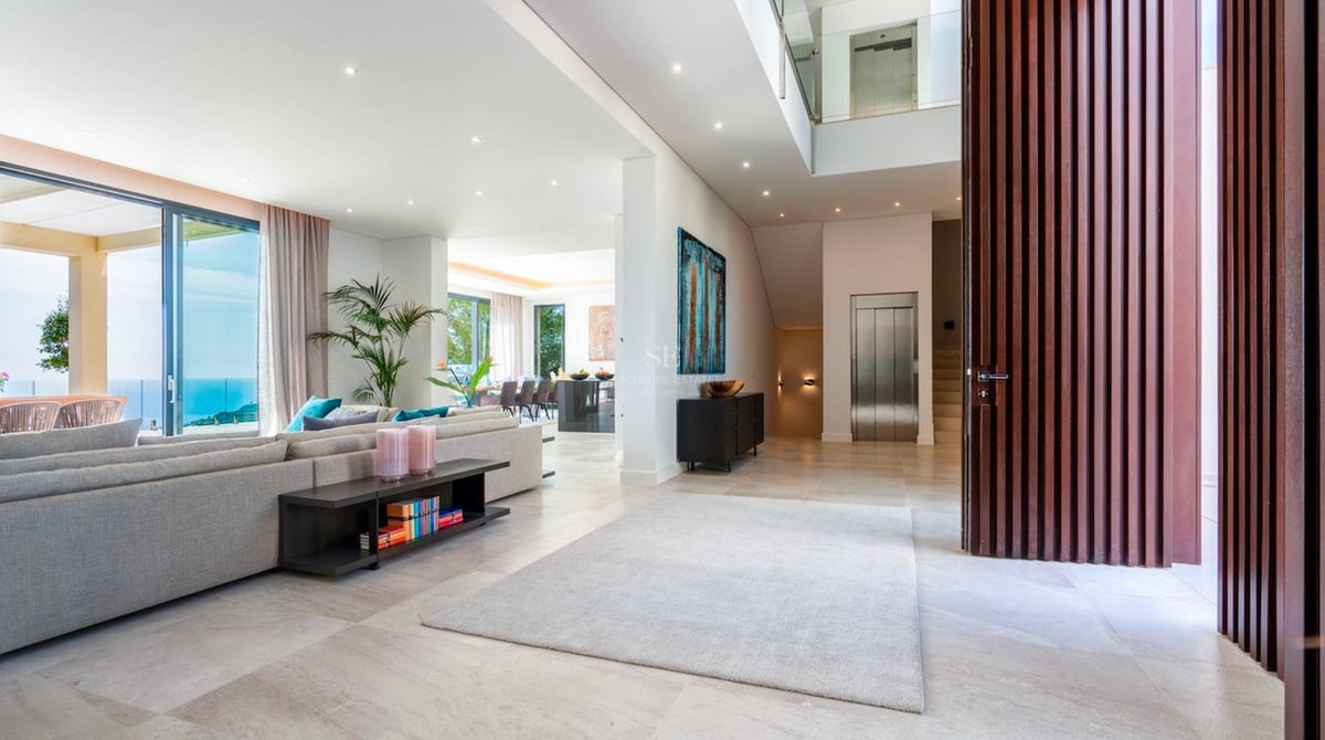 Salon moderne spacieux avec canapé gris, grandes baies vitrées avec vue sur mer et ascenseur intérieur.