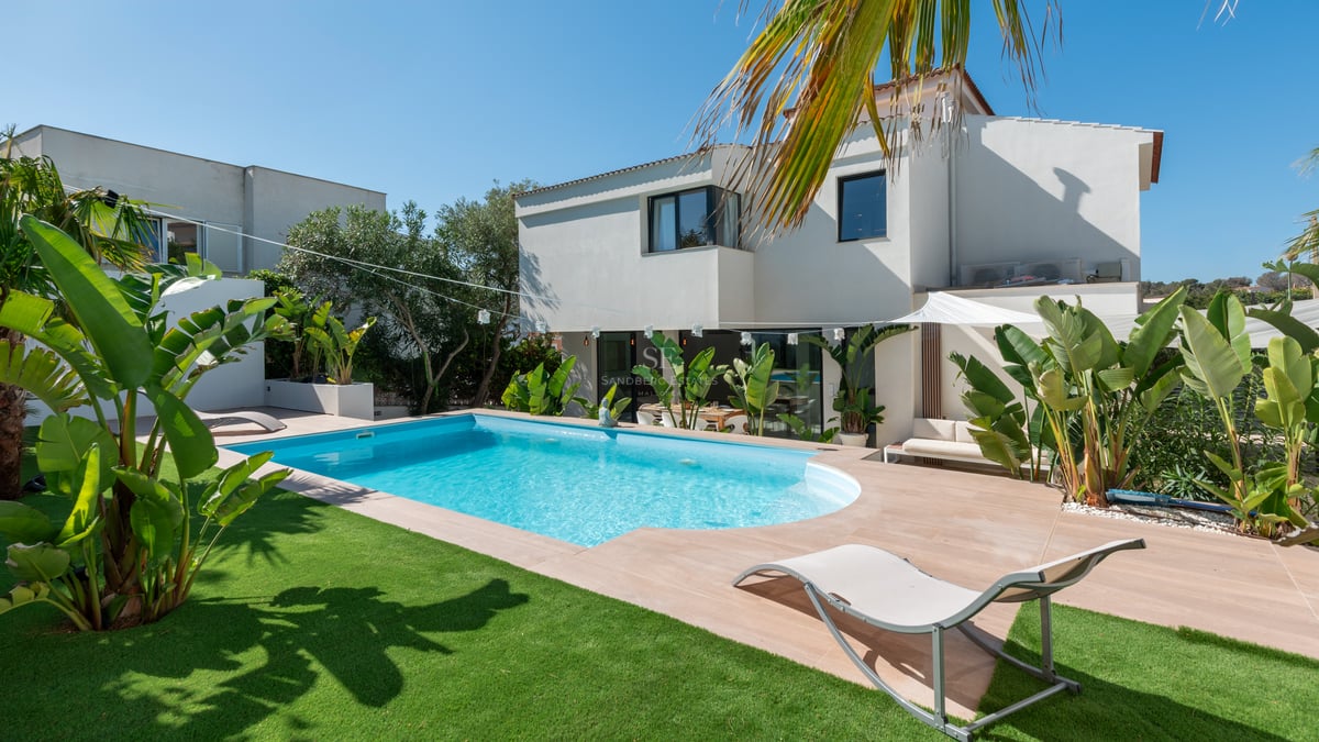 Villa blanche moderne avec piscine bleue, transat, pelouse verte et plantes tropicales par temps clair.