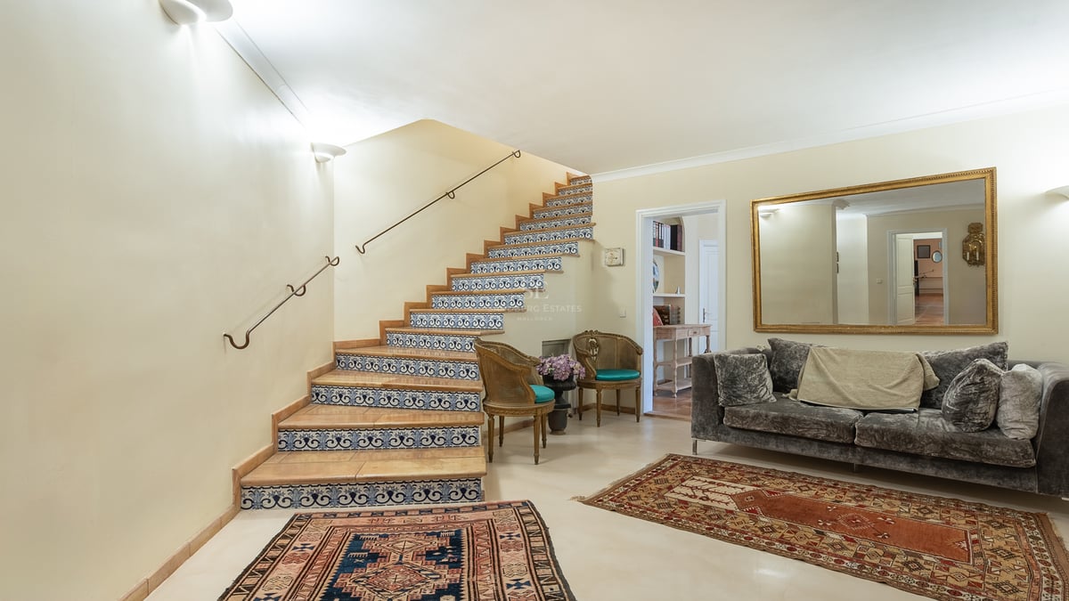 Interior view of an entrance hall featuring a staircase with blue and white tiles, a velvet sofa, and oriental rugs.