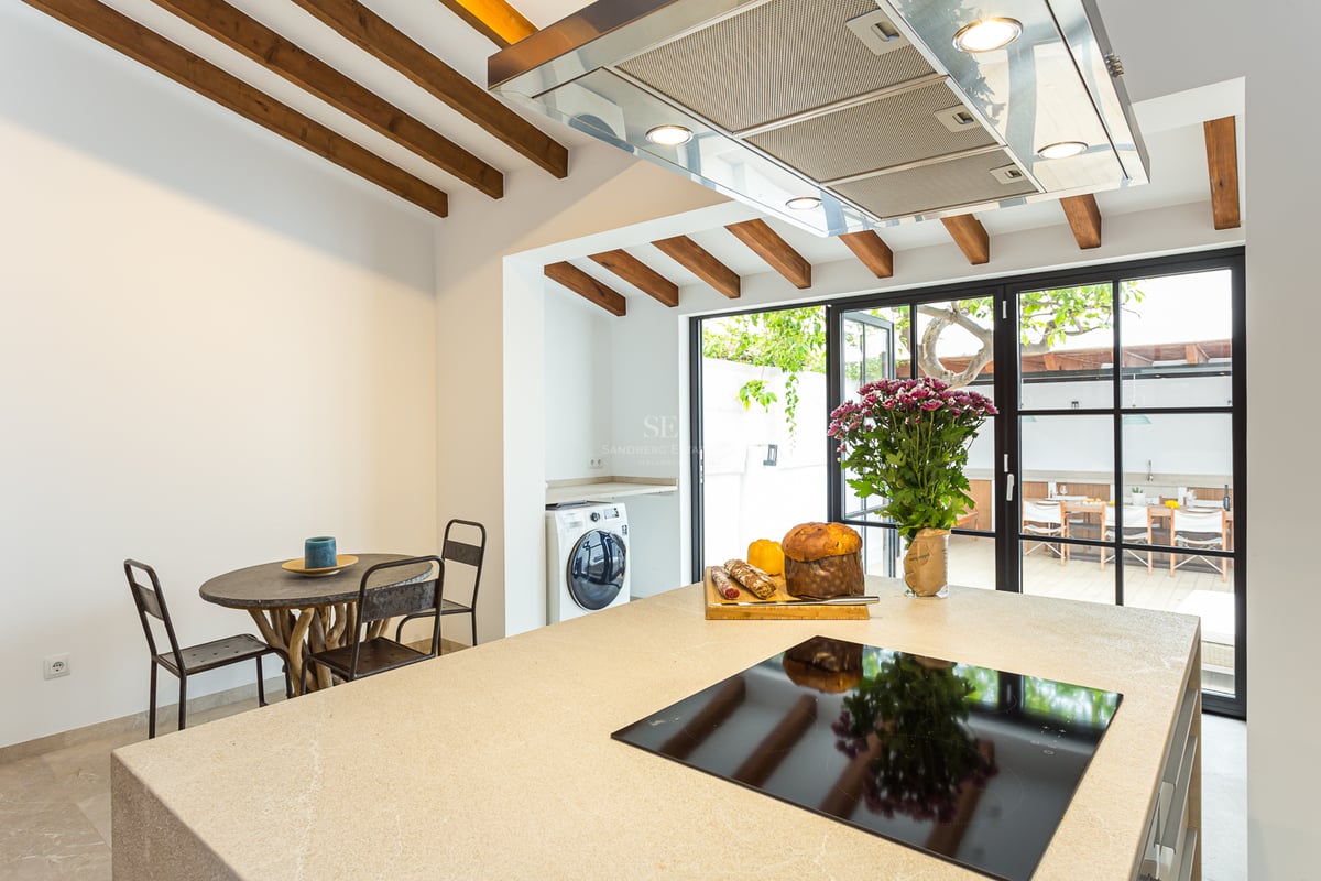Une cuisine moderne avec un îlot en pierre beige, plaque à induction et poutres rustiques donnant sur une terrasse.
