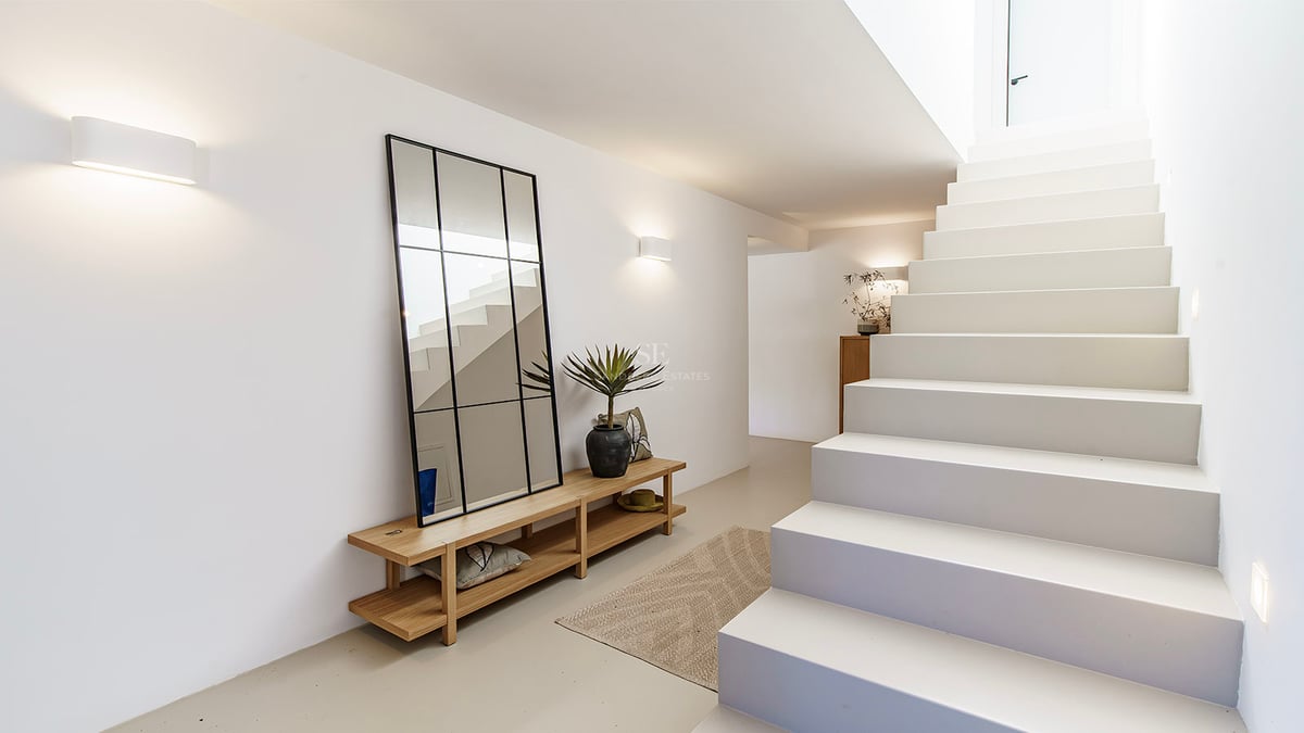 Couloir minimaliste lumineux avec escalier blanc, grand miroir à cadre noir, banc en bois et éclairage mural chaleureux.