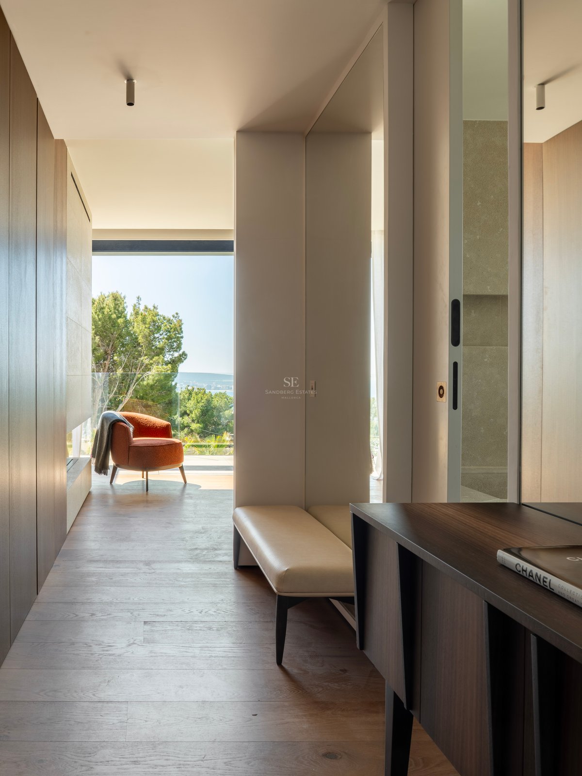 Couloir moderne, parquet en bois, fauteuil terracotta et grandes baies vitrées offrant une vue sur mer et pins.