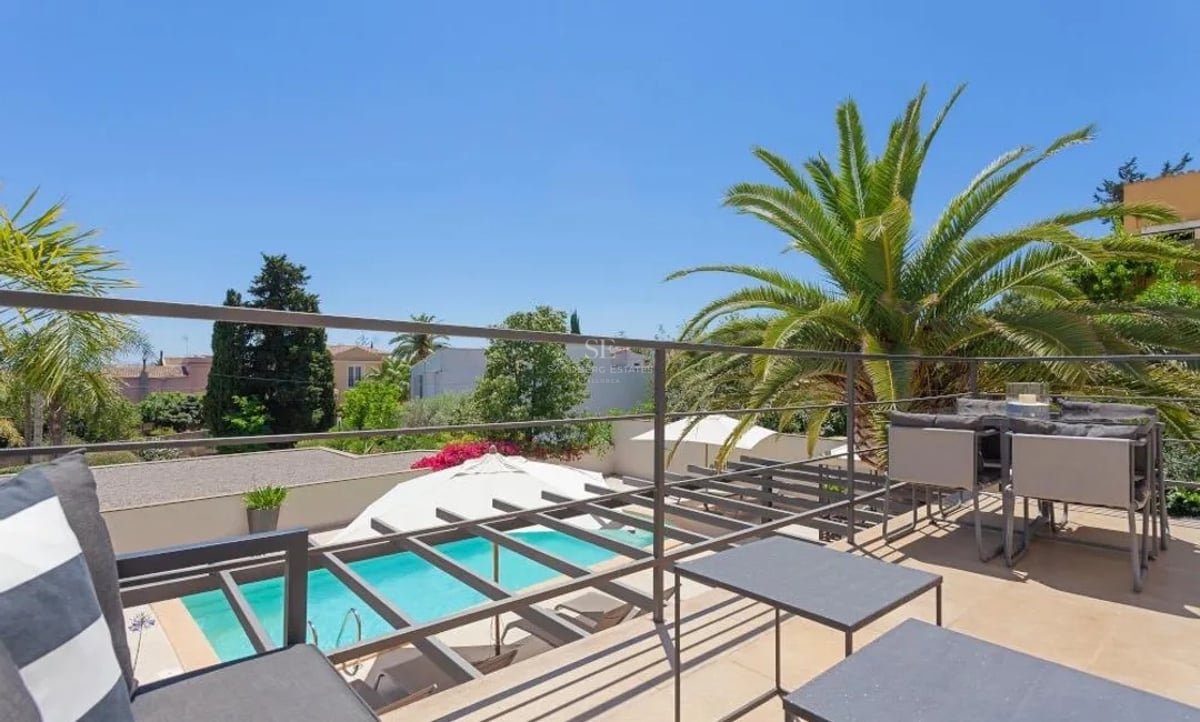 Terrasse surélevée avec mobilier en métal gris, vue sur une piscine turquoise et des palmiers sous un ciel bleu azur.