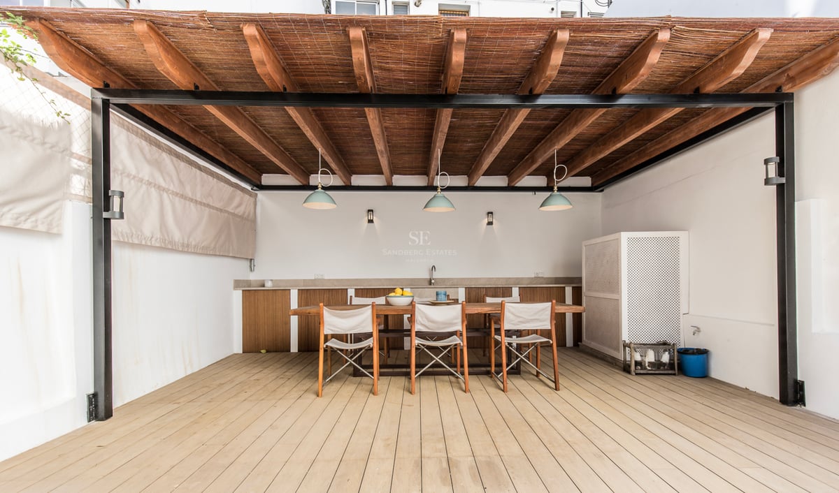 Salle à manger extérieure ombragée avec une grande table en bois, des chaises et une pergola avec kitchenette.