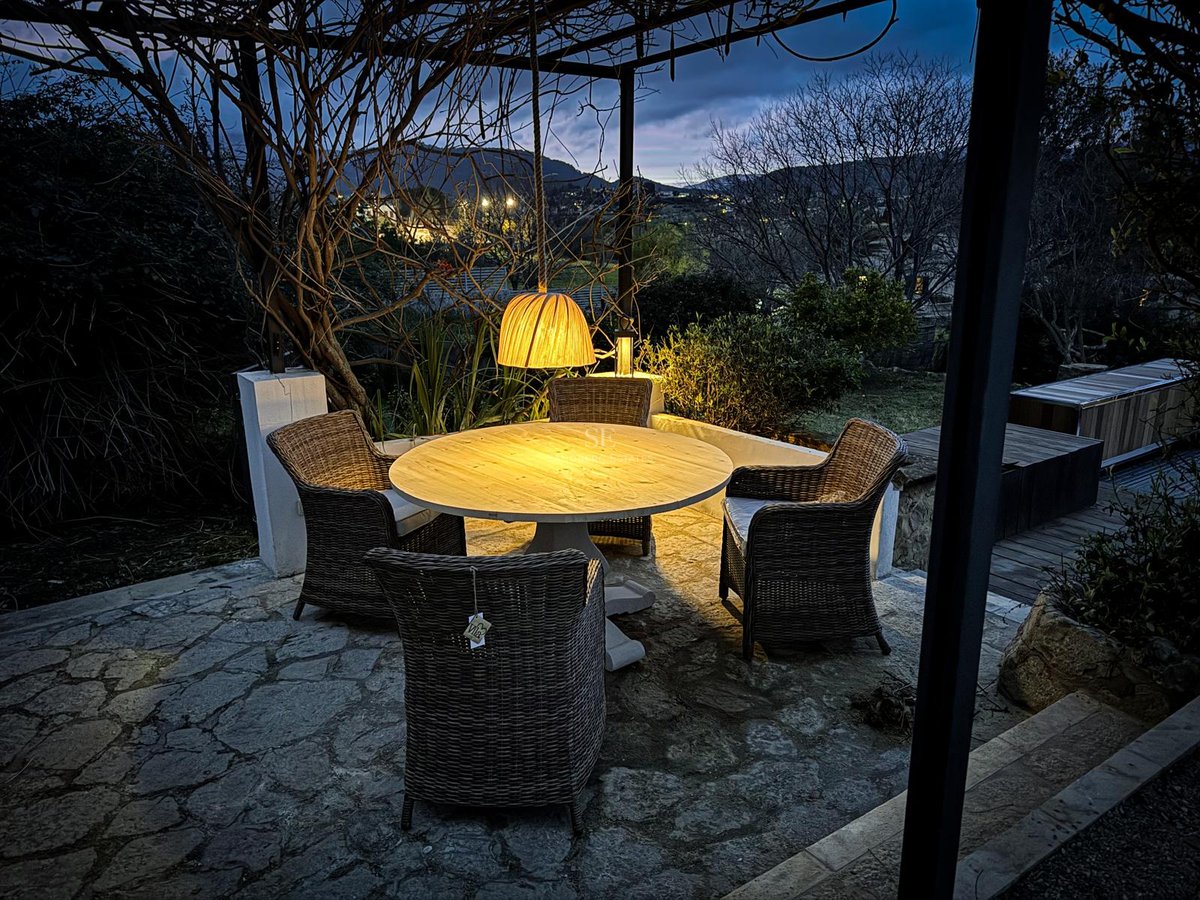 Terrasse extérieure en pierre avec table en bois, chaises en osier et lampe chaleureuse surplombant les collines.
