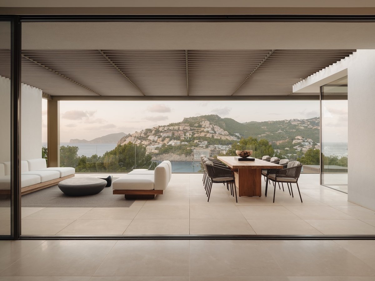 Terrasse couverte moderne avec table en bois, canapés blancs et vue sur mer à travers des garde-corps en verre.