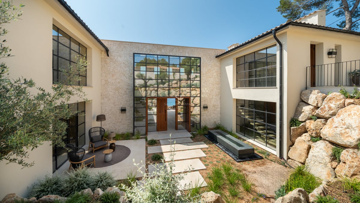 Entrée de villa moderne avec façade en pierre, grandes fenêtres à carreaux et patio paysager avec salon extérieur.