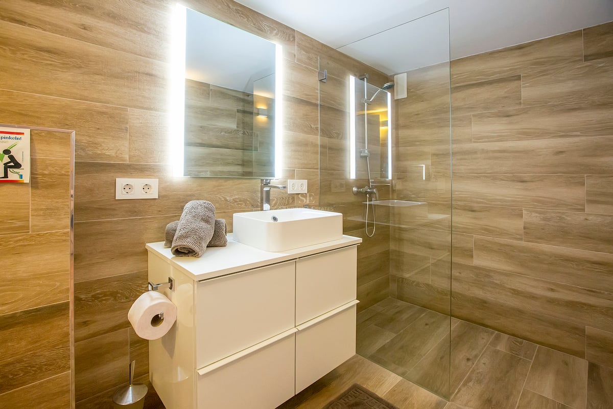 Salle de bains contemporaine avec carrelage effet bois, douche à l'italienne et vasque blanche avec miroir rétroéclairé.