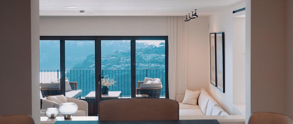 Salon lumineux avec canapé blanc et grandes baies vitrées donnant sur une colline et du mobilier de balcon.