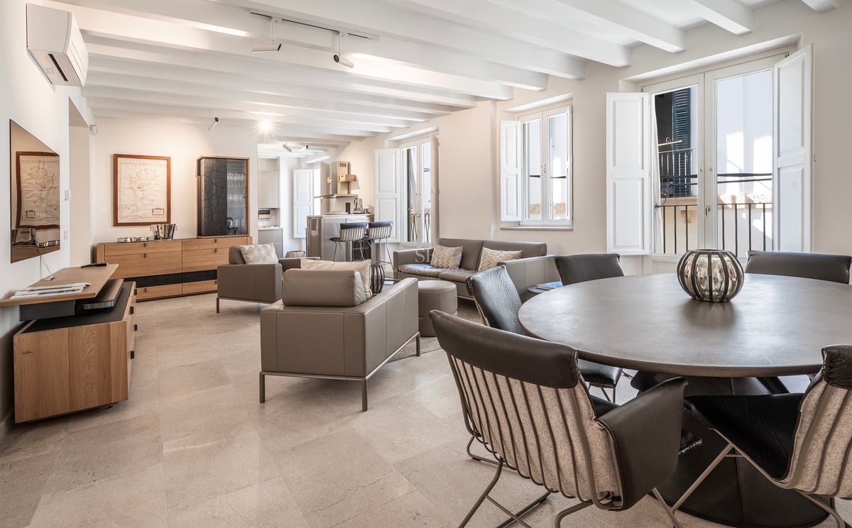 Salon spacieux avec plafonds à poutres blanches, meubles en cuir gris et table à manger ronde.