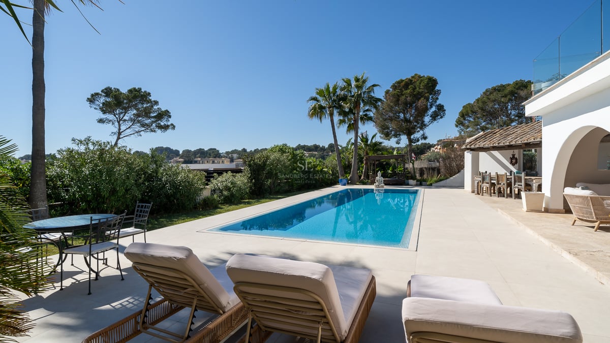Piscine turquoise entourée de dalles blanches, chaises longues, palmiers et villa méditerranéenne sous un ciel bleu.