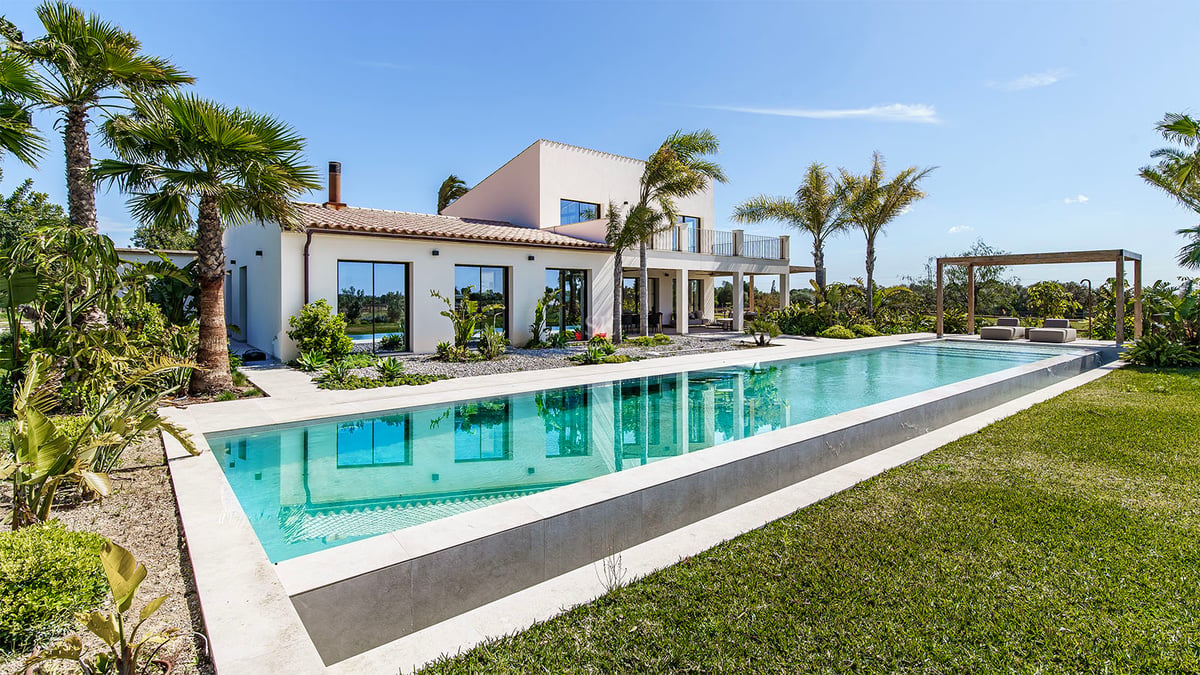Villa blanche contemporaine avec une grande piscine rectangulaire, pelouse soignée et palmiers sous un ciel bleu clair.