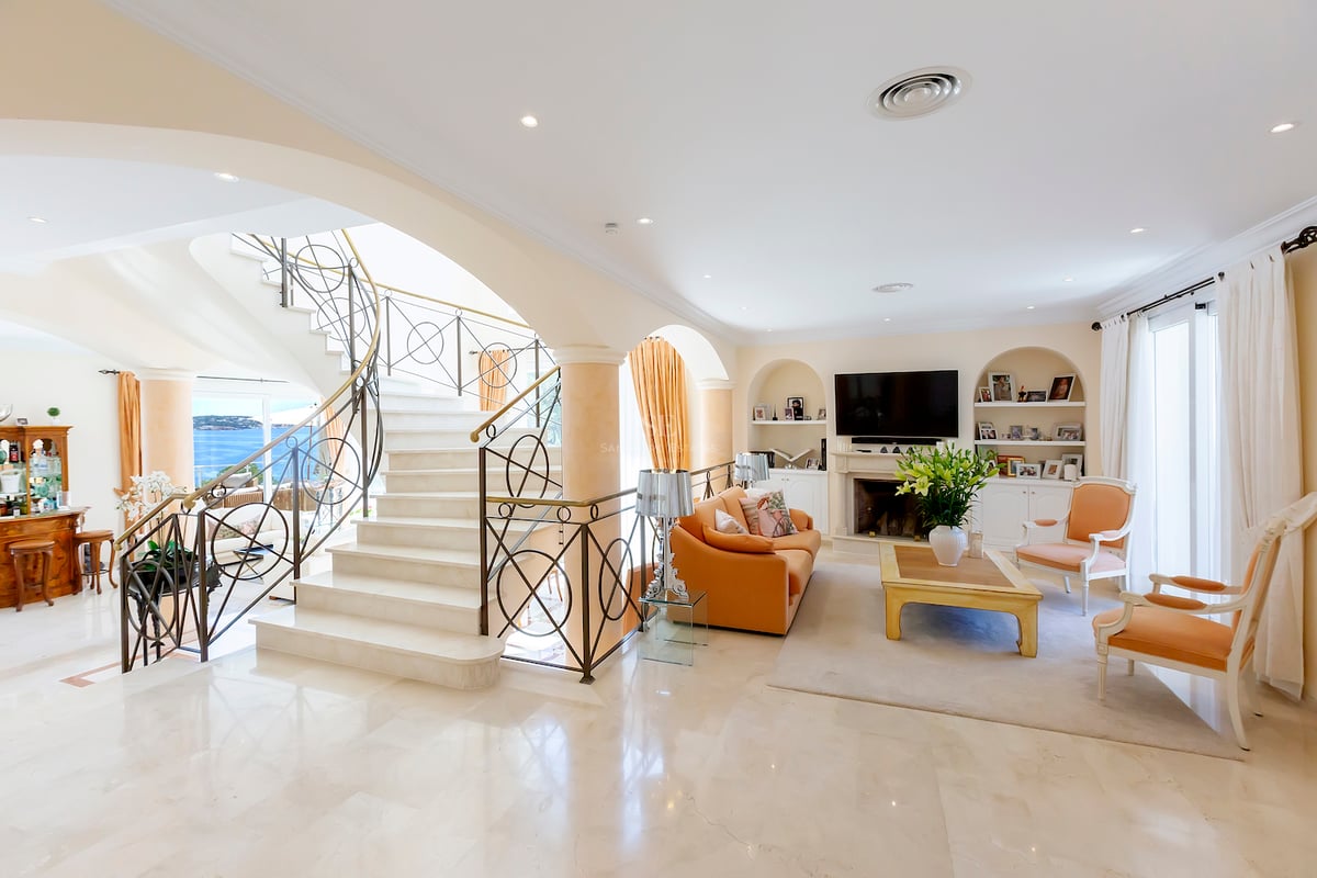 Salon spacieux avec grand escalier en marbre, fenêtres cintrées avec vue mer et cheminée classique.