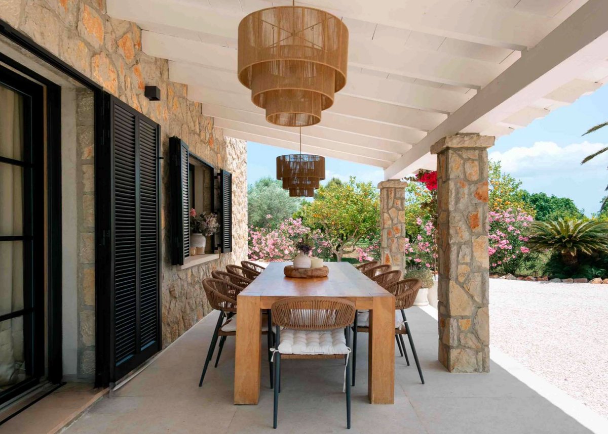 Grande table en bois sous un porche avec colonnes en pierre, luminaires en rotin et vue sur un jardin fleuri.