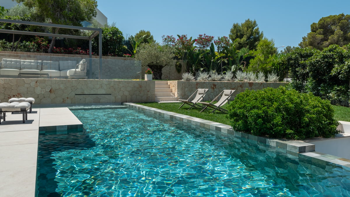 Piscine moderne avec eau turquoise, murs en pierre naturelle et jardin paysager sous un ciel bleu.