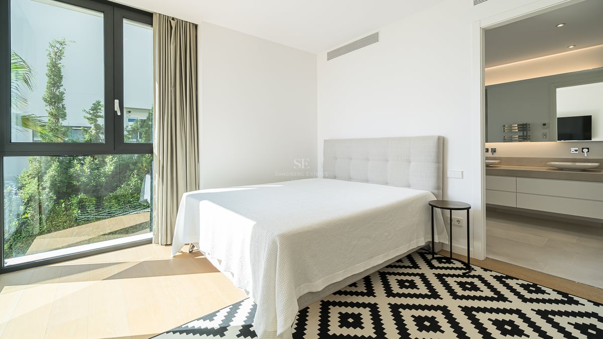 Chambre moderne avec lit blanc, tapis géométrique, grande fenêtre sur jardin et salle de bain attenante.