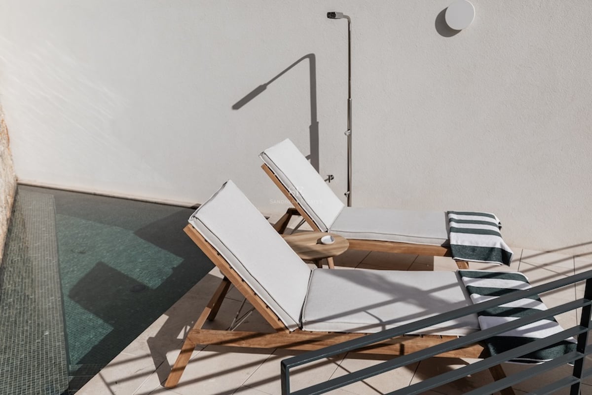 Deux chaises longues en bois avec coussins blancs à côté d'une piscine et d'une douche extérieure sur une terrasse ensoleillée.