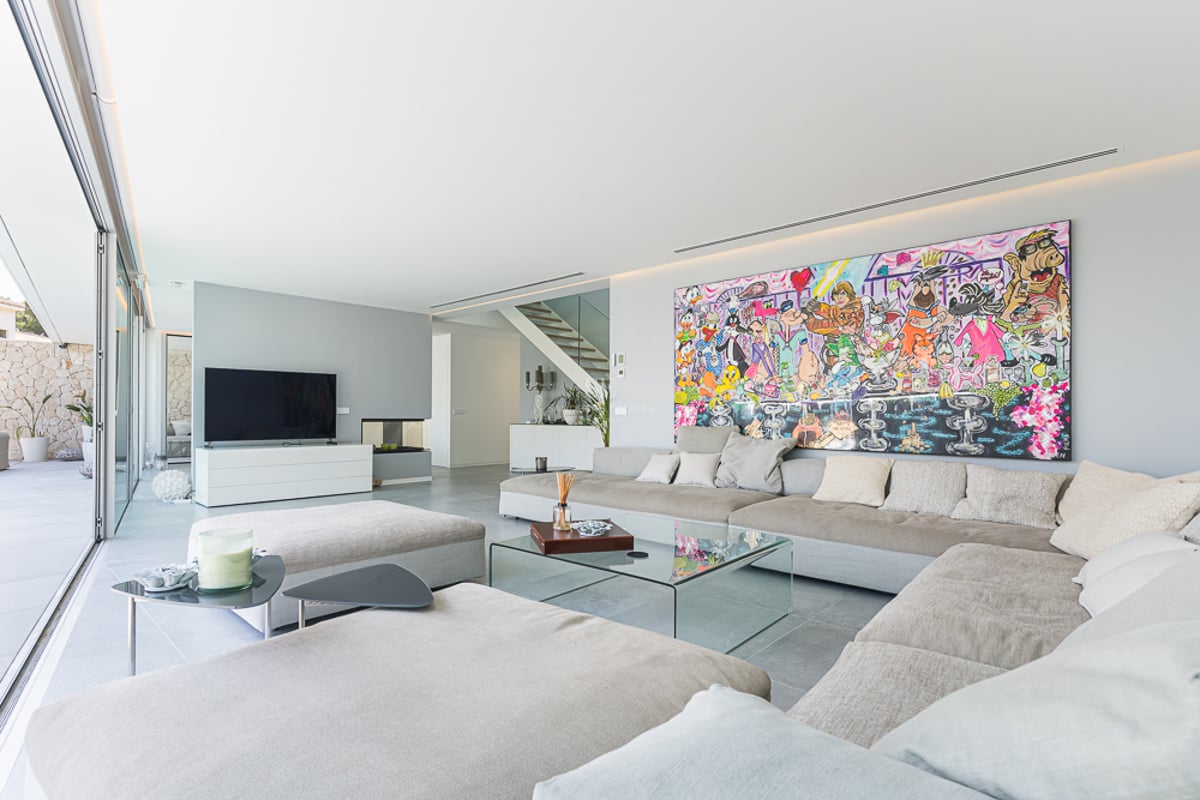 Salon moderne avec canapé beige, table en verre et un grand tableau coloré sur un mur blanc.