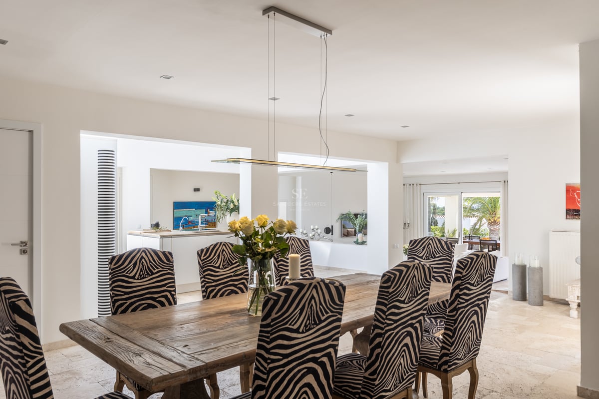 Table à manger en bois rustique, chaises à motif zébré et sols en travertin dans une villa lumineuse.