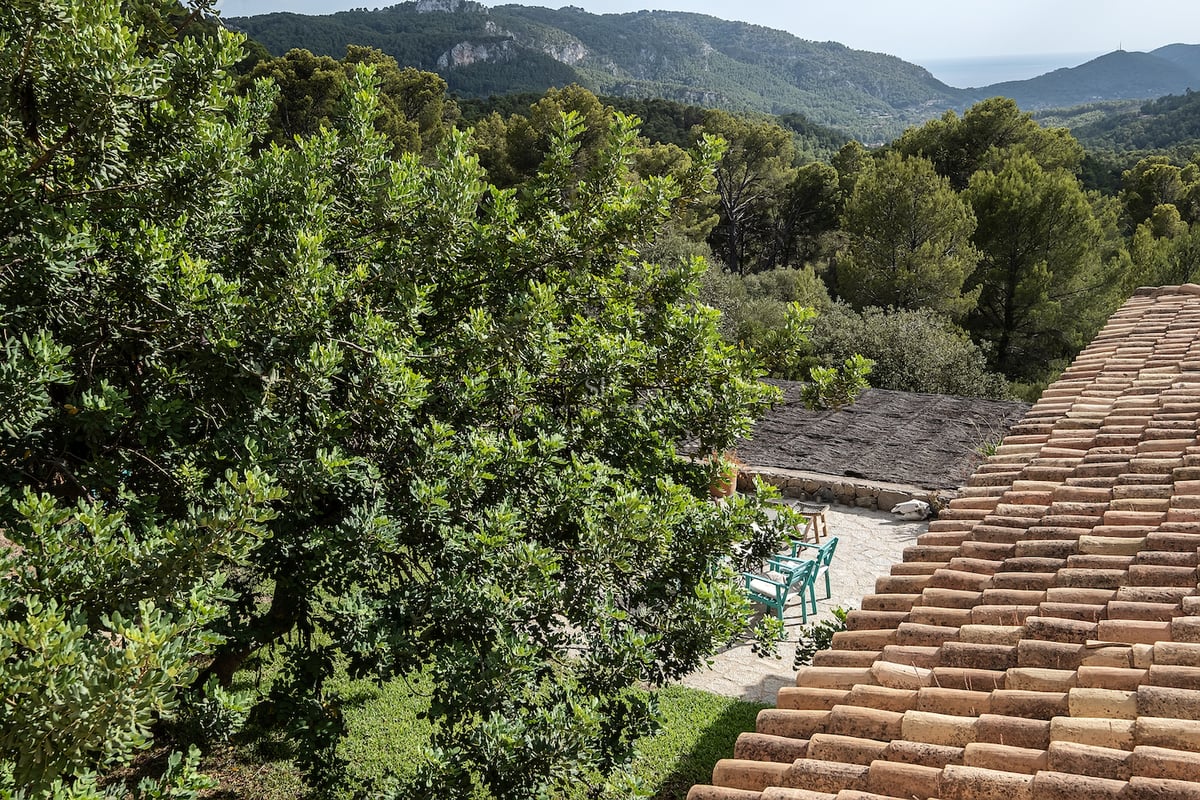 Toit en terre cuite surplombant un patio en pierre avec des chaises turquoise, entouré d'arbres et de montagnes.