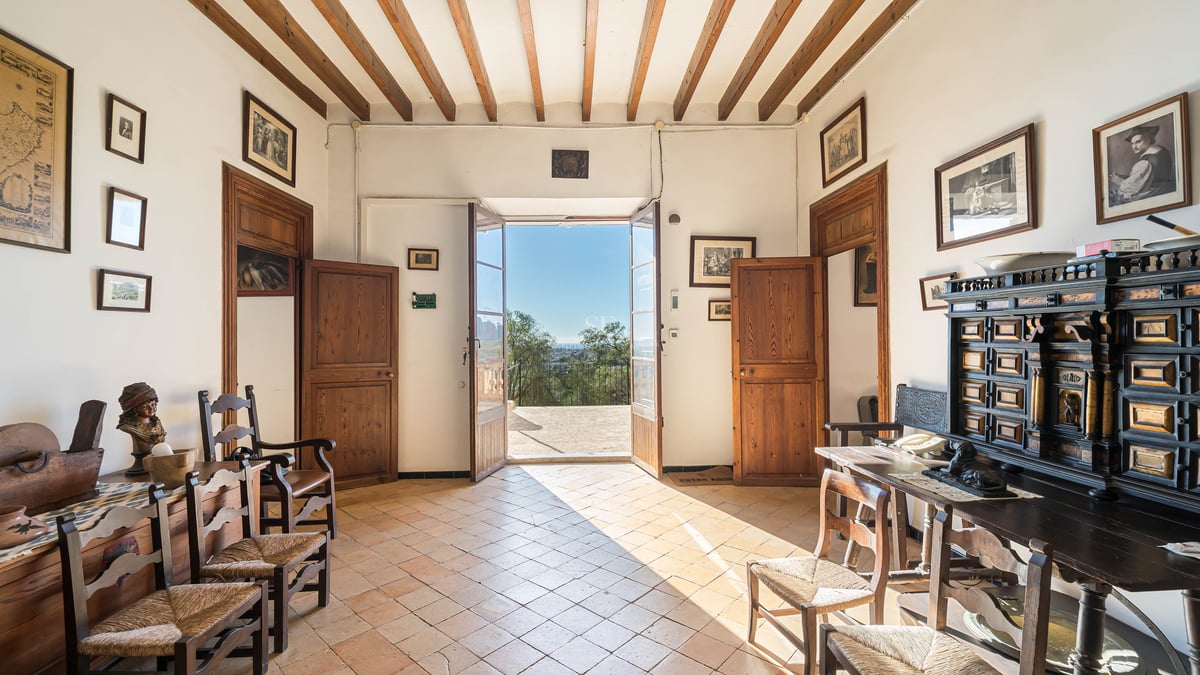 Hall traditionnel avec poutres en bois, sol en terre cuite et portes ouvertes donnant sur une terrasse ensoleillée.