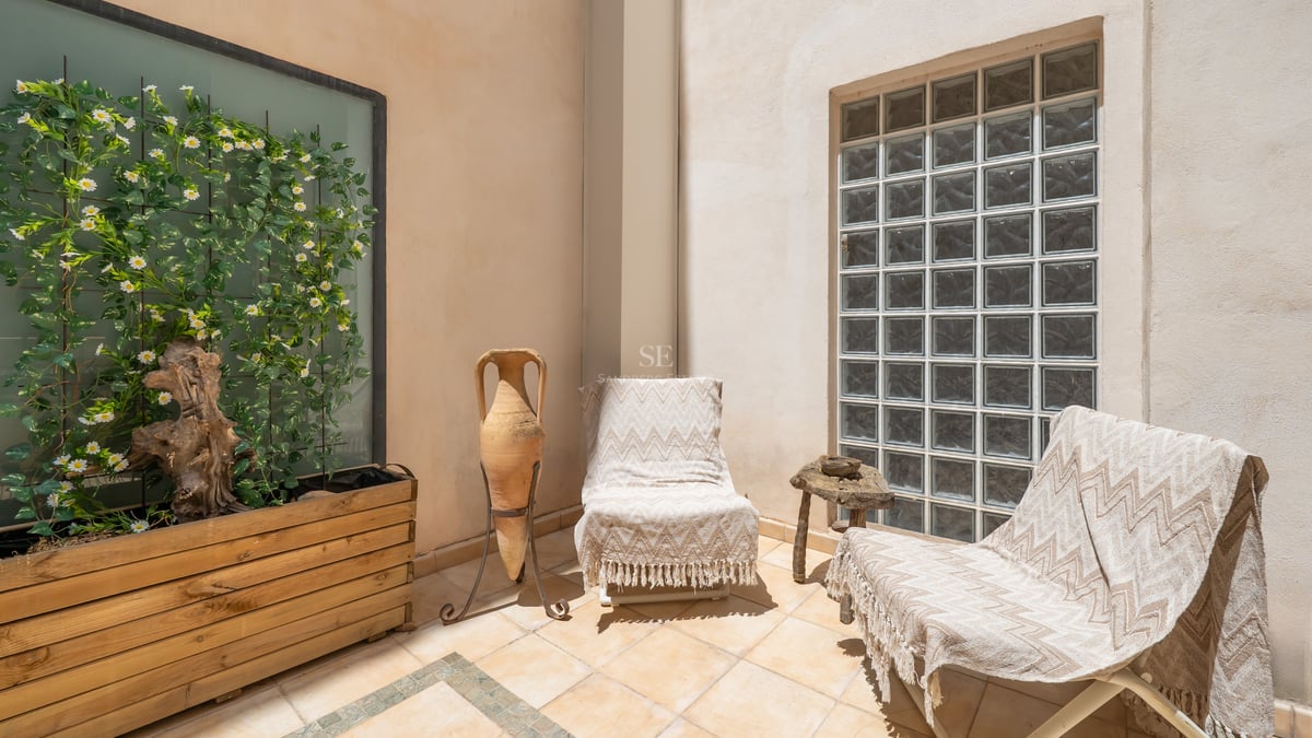 Patio ensoleillé avec deux chaises longues, une amphore en terre cuite et un mur en briques de verre.
