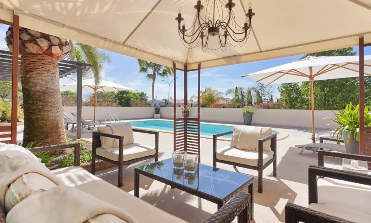 Terrasse ombragée avec mobilier en rotin et lustre, vue sur une piscine privée et des palmiers sous un ciel bleu.