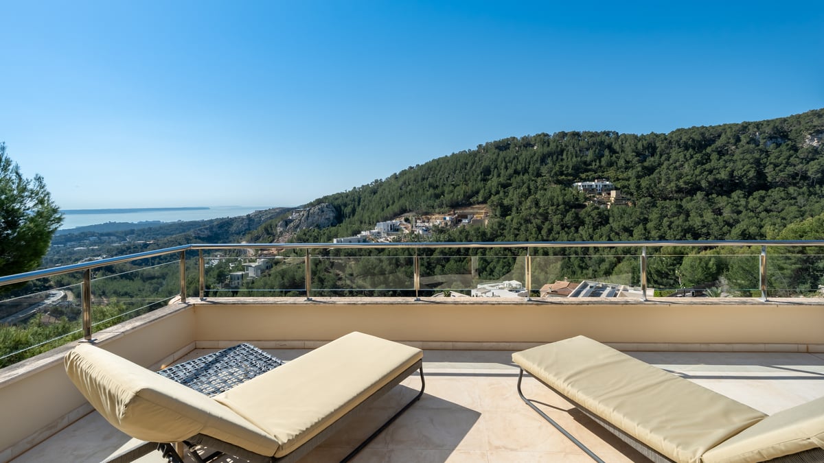 Deux chaises longues crème sur une terrasse en pierre avec garde-corps en verre surplombant une vallée et la mer.