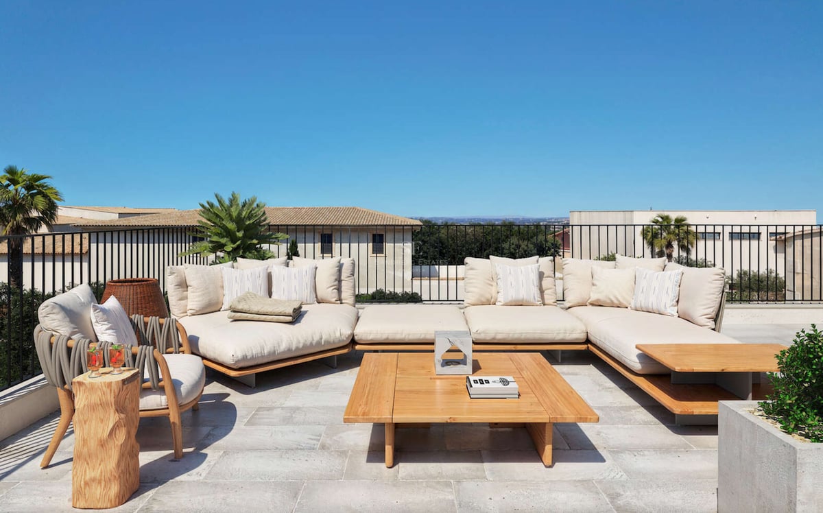 Terrasse ensoleillée avec grand canapé d'angle crème, table basse en bois et sol en pierre naturelle.