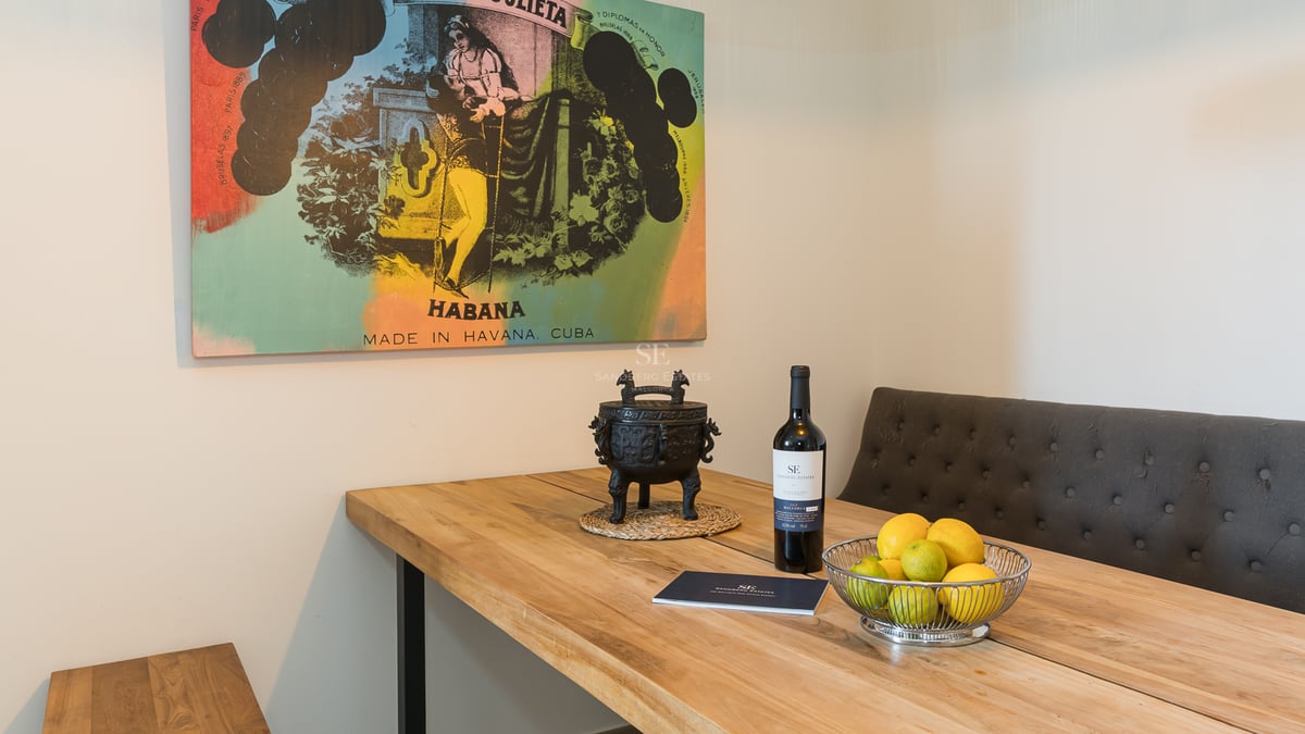 Table à manger en bois massif avec banc rembourré gris, bouteille de vin, bol de citrons et tableau vintage cubain.