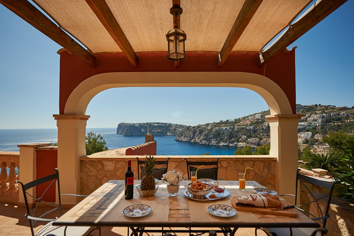 Une terrasse couverte avec une table dressée pour le petit-déjeuner donnant sur une baie bleue et des falaises côtières.