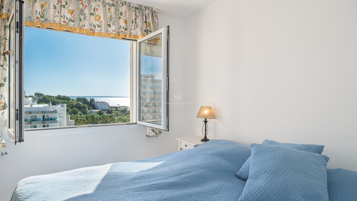 Chambre avec lit bleu et fenêtre ouverte offrant une vue sur un paysage côtier avec mer et arbres.